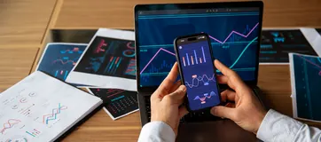 A hand holding a phone with an investment app in front of a tablet showing market charts