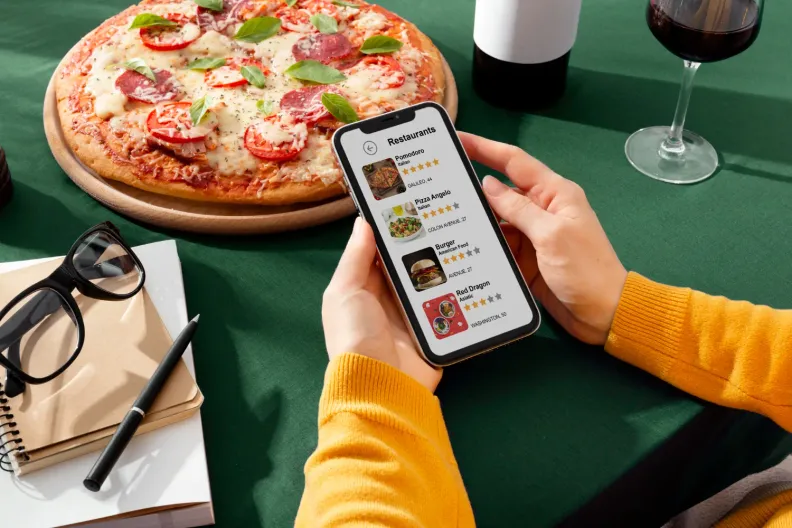 Person browsing a restaurant list on a phone beside a pizza